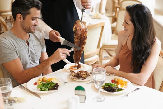 Happy couple, date and waiter cutting kebab for serving, fine dinning or romantic dinner at table. Young man and woman with chef or skewer of food for meal, eating or enjoying service at restaurant