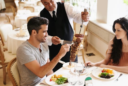 Happy couple, date and waiter with kebab for serving, fine dinning or romantic dinner at table. Young man and woman with chef cutting skewer of food for meal, eating or enjoying service at restaurant