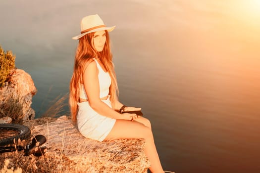 A tourist woman is sitting by the sea in a hat and white summer clothes, looking happy and relaxed.