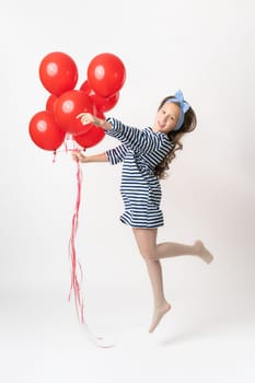 Playful girl in striped dress is jumping, holding bunch of red balloons in hand, looking at camera