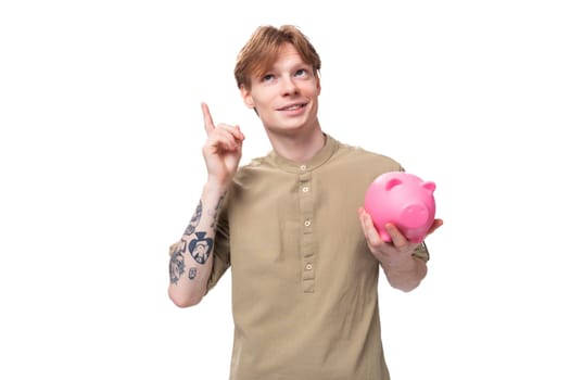 young handsome red-haired man in glasses and a shirt keeps savings in a piggy bank on a white background