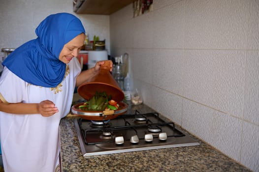Smiling Muslim woman in blue hijab, opening lid of a clay dish while cooking veggies in Moroccan tagine at home kitchen