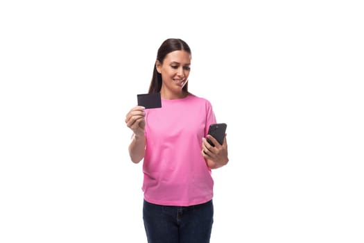 young slim brunette woman in pink basic t-shirt holding credit card mockup and smartphone on white background with copy space