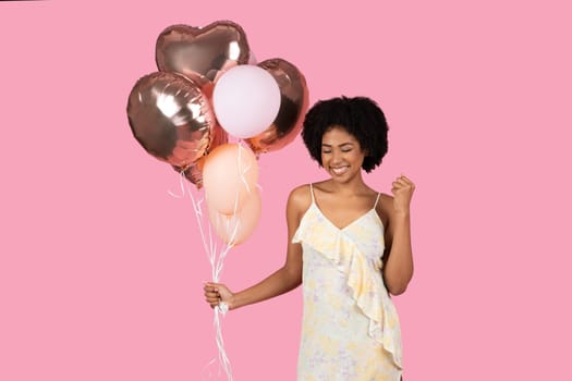 Joyful African American woman with curly hair, playfully holding a cluster of rose gold balloons