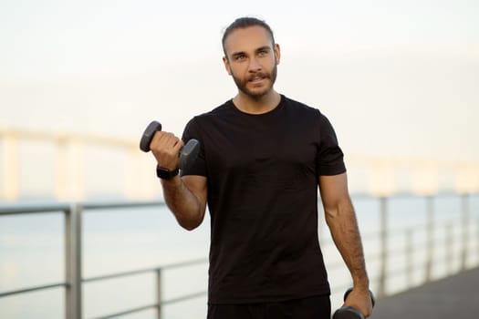 Muscular man lifting heavy dumbbells exercising at sea pier outdoor