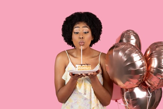 Delighted African American woman with curly hair blowing out a candle on a slice of birthday cake