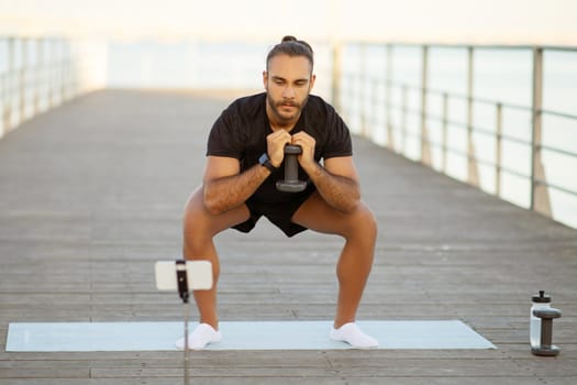 fitness blogger guy squats with dumbbells on a sea pier