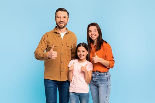Family of three gesturing thumbs up smiling at camera in studio over blue background, caucasian parents and their little preteen daughter approving offer with like symbol