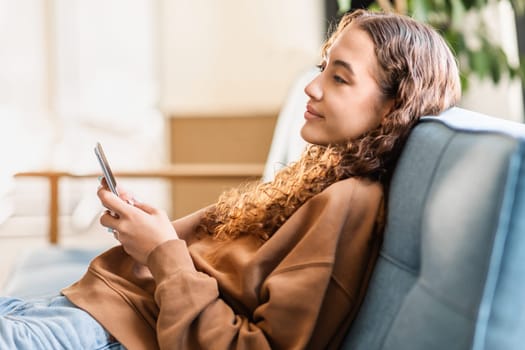 Adolescent European girl holding cellphone resting on sofa at home