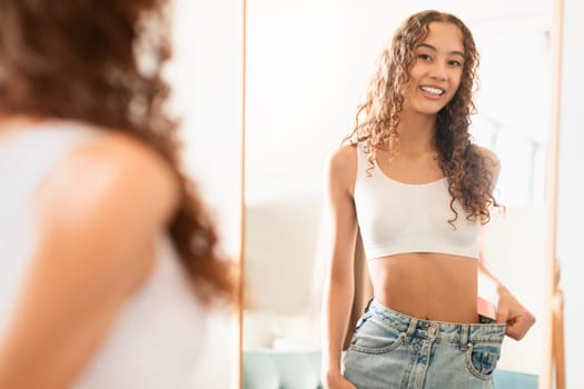 Caucasian teenager girl at mirror showcasing weight loss success indoors