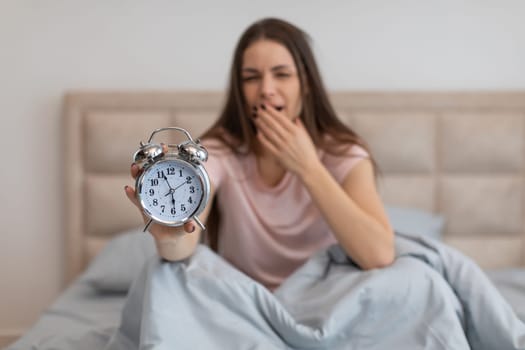 Tired woman in bed with alarm clock, yawning early in the morning