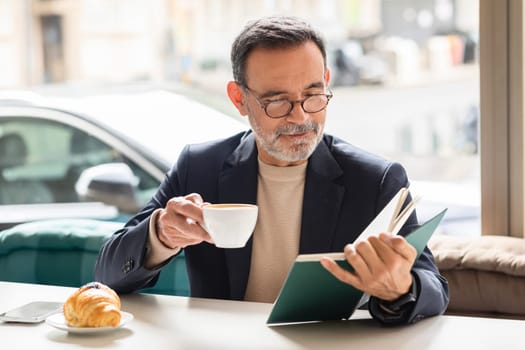 Intelligent mature man with glasses enjoying a cup of coffee while reading a book