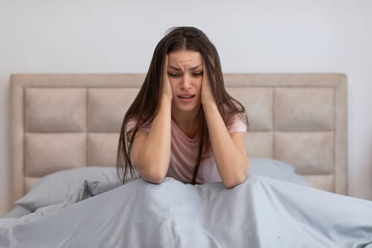 Distressed woman sitting in bed, holding her head, showing signs of headache or intense stress