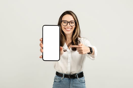 Engaging young woman showcasing a smartphone with a pointing gesture, wearing glasses