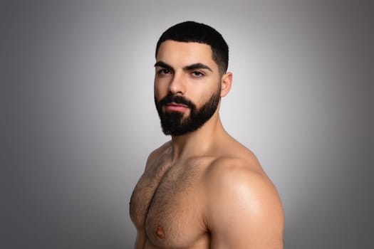 Young Middle Eastern guy with beard, shirtless, posing on grey