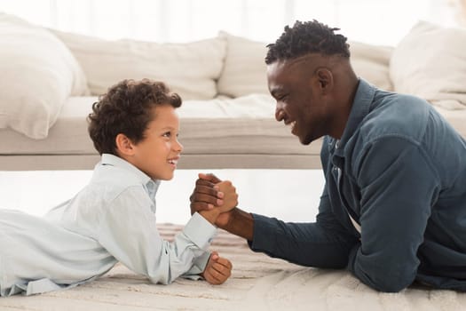 Happy African American Daddy And Little Son Arm Wrestling Lying On Floor At Home. Happy Young Daddy And Little Boy Competing And Having Fun While Armwrestling Together. Side View