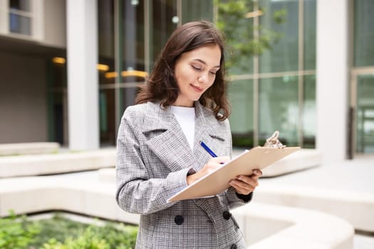 Young Woman Holding Clipboard Reading And Signing Business Papers Outside