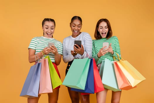Amazed three young multiethnic ladies shopping online, using smartphones