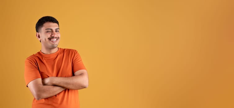Positive brazilian man in orange t-shirt posing with crossed arms on chest, winking and smiling, panorama, free space