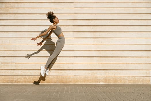 Female athlete in sportswear leaping in air with wall backdrop outdoors. Healthy lifestyle concept