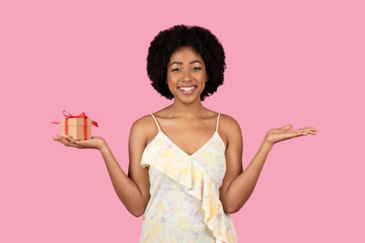 Radiant African American woman with curly hair, smiling and balancing a small gift box with a red ribbon