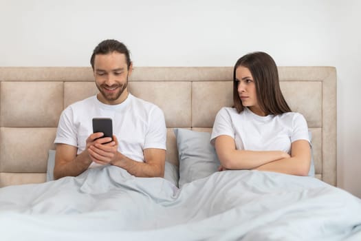 Man is happily engaged with his smartphone while the woman next to him looks on with concerned and displeased expression, hinting at digital distraction