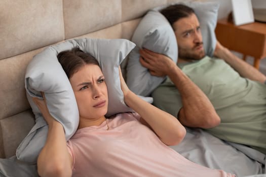 Disturbed couple covering ears with pillows in bed