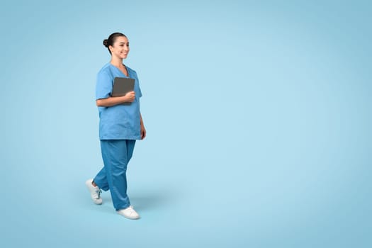 Cheerful young female nurse in scrubs holding tablet while walking, looking positive and professional, blue background, free space, medical banner