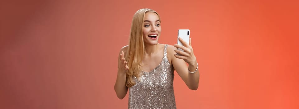 Charming elegant nice blond girl in silver dress talking video call speaking looking smartphone display amused surprised smiling happily have conversation sibling showing prom outfit.