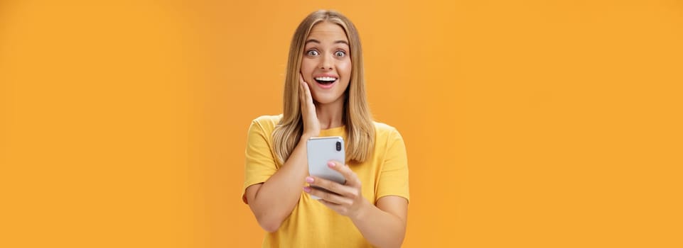 Portrait of surprised and impressed woman reacting to awesome app in smartphone touching cheek from amazement and joy smiling broadly at camera holding cellphone in hand over orange wall. Technology and emotions concept
