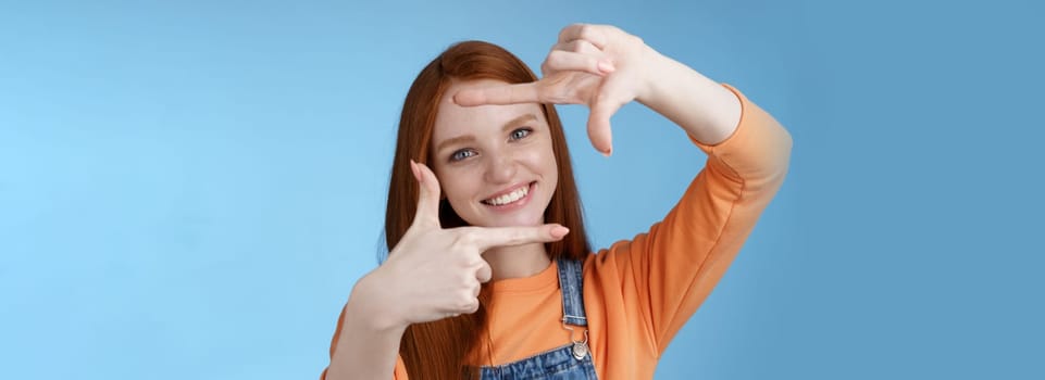 Joyful attractive sincere redhead young girl searching inspiration find perfect angle take good shot make hand frames look through delighted amused smiling broadly white teeth, blue background.
