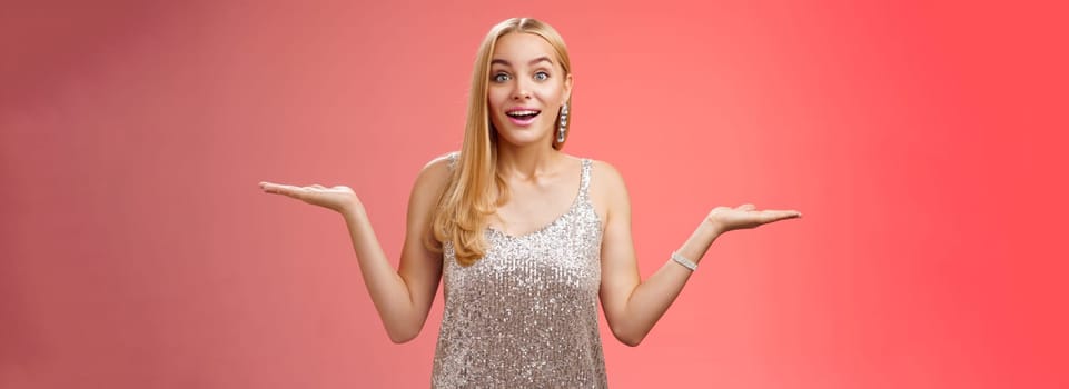 Carefree fascinated young blond girl 20s in silver glittering evening party dress raising hands sideways weighing shrugging smiling amused search right choice, deciding what do red background.