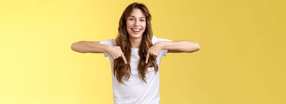 Cheerful lively happy funny girl long beautiful hair pointing down index fingers indicate bottom promo laughing joyfully share positive emotions recommend product advertising yellow background.