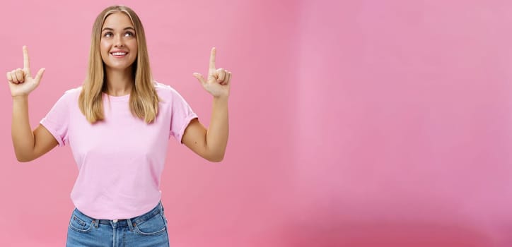 Girl daydreaming pointing and looking with upbeat and admiring expression up smiling cheerfully enjoying nice sunny weather walking in casual t-shirt and jeans posing against pink background. Copy space
