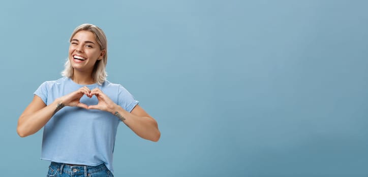 Love relationship and emotions concept. Joyful charming feminine girl with fair hair in trendy t-shirt winking happily smiling broadly and making heart gesture over breast expressing affection.