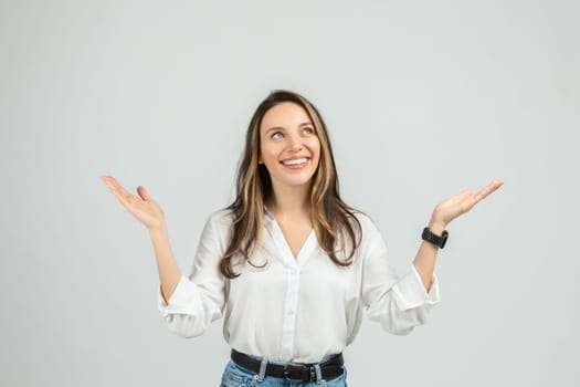 An upbeat young woman in a white blouse and blue jeans is raising her hands, palms up