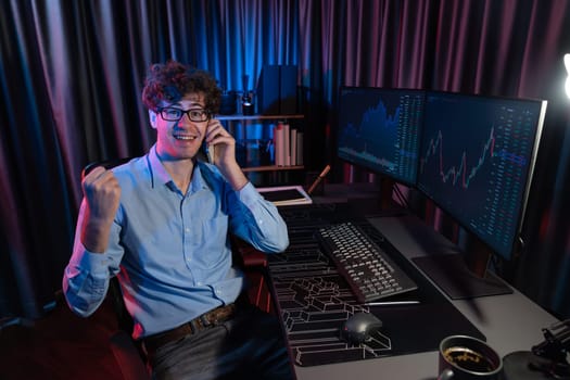 Successful young business trader raising fist up with highest profit stock with looking at camera in real time market data graph at neon office, calling to broker earning valued rate upper. Gusher.