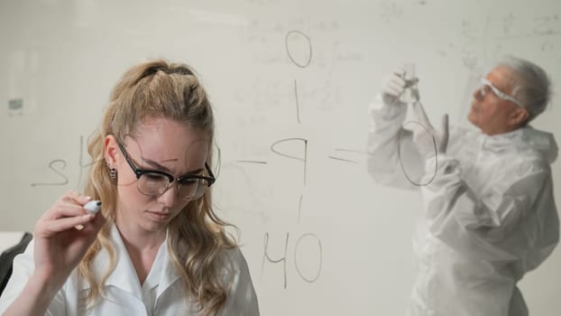 A woman chemist writes a formula on glass. An elderly Caucasian man in a protective suit is doing tests.