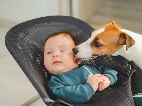 A dog licks the face of a cute three-month-old boy dressed in blue overalls in a baby lounger.