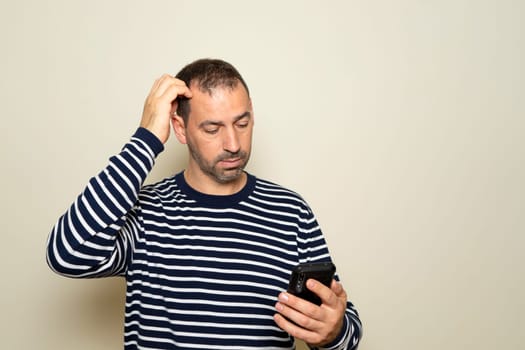 Bearded Hispanic man in his 40s wearing a striped sweater scratching his head while searching for the solution to a problem on his smartphone isolated over beige background.