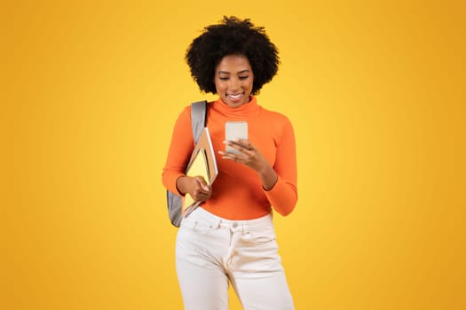 Engaged young woman with afro hair, wearing an orange sweater, looking at her smartphone with a smile