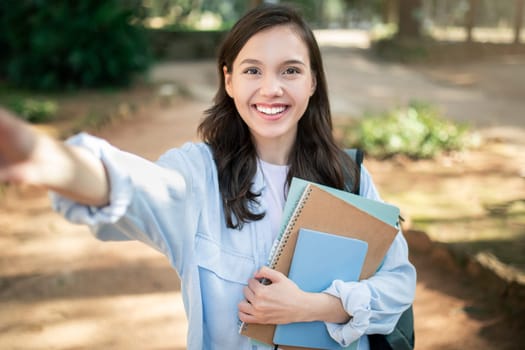 A radiant young woman smiles at the camera, taking a selfie while clutching notebooks on a sunny outdoor campus