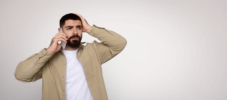 Worried millennial arab man in a khaki shirt and white t-shirt talks on the phone while anxiously touching his head, expressing concern or confusion during the conversation