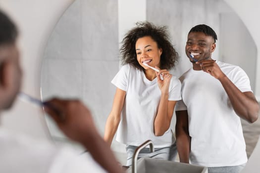 Smiling black married couple enjoys morning dental routine, brushing teeth together for tooth care and shared wellness in their home bathroom. Selective focus on mirror reflection