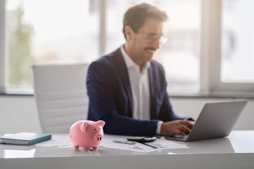 Smiling middle aged european businessman in a blue suit works on a laptop at a white desk with a pink piggy bank in the foreground, symbolizing financial planning. Work, business