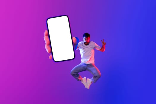 Joyful Caucasian guy holding big smartphone with empty screen, jumping in studio lit with purple and neon lights, advertising wow mobile applications and online communication offers