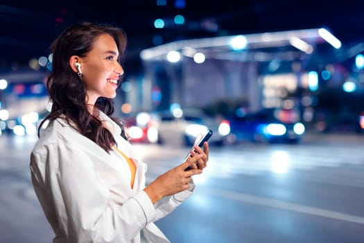 Young happy lady with earphones, standing on city street at night, listening music from smartphone while walking, illuminated city night lights on background, side view, copy space