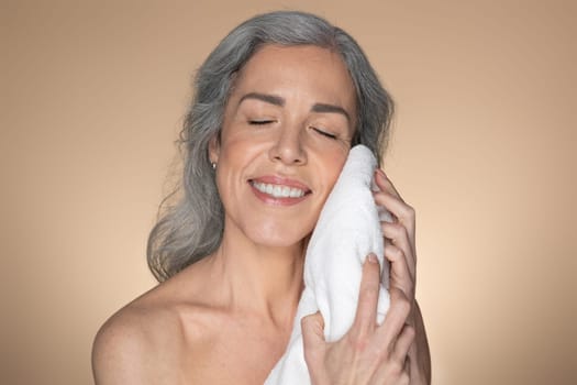Happy caucasian senior woman wiping face with soft towel and smiling, standing over beige studio background