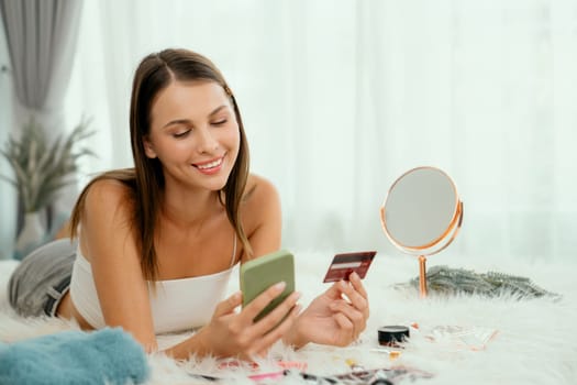 Young woman lying on the bed using online payment app and digital wallet on smartphone to pay with credit card. E commerce shopping and modern purchasing via mobile internet. Unveiling
