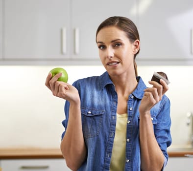 Woman, portrait and choice with apple, cupcake and decision in health and wellness at home. Face of female person with organic green fruit and chocolate muffin for diet or option in kitchen at house